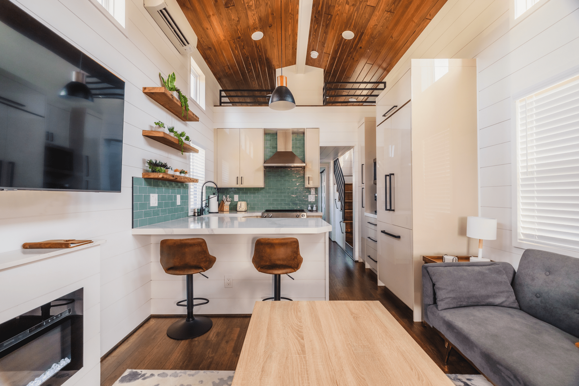 View of a Black prong resort cabin showing the barstool kitchen, wood slatted ceiling with an upstairs loft and a cozy couch and mounted TV in the foregrond - By InfinityPhotos